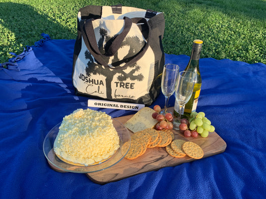 The image shows an ivory picnic bag with black handles. On the front of the bag is a black silhouette of a Joshua tree, with the words "JOSHUA TREE" and "CALIFORNIA" written below it. Our Picnic Bag is READY for ROMANCE! Large Enough to Carry A Full-Size Bottle of Champagne or Wine and Drinking Glasses as well as Food and a Picnic Blanket for those Last-minute Spontaneous GetAways!