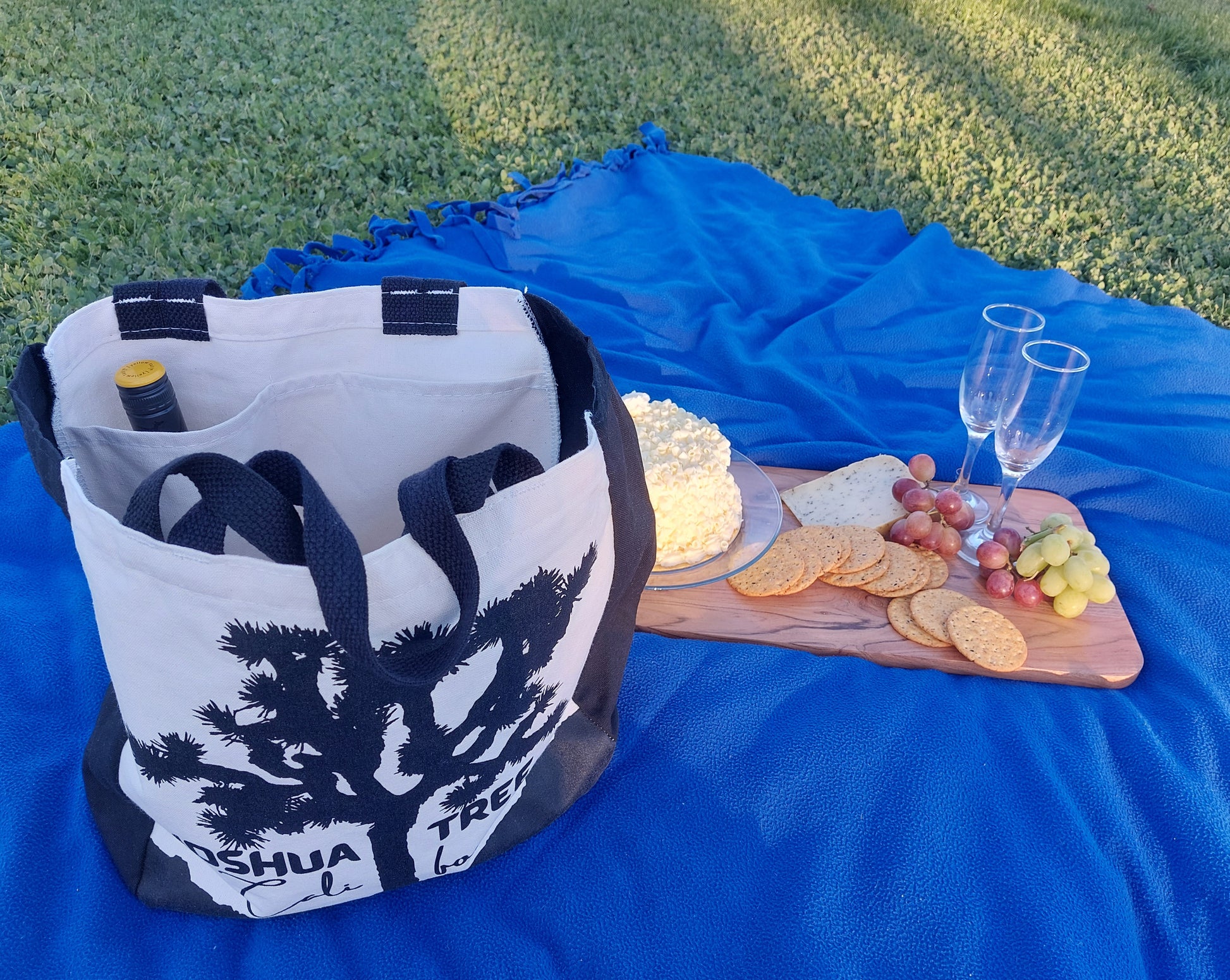 The image shows an ivory picnic bag with black handles. On the front of the bag is a black silhouette of a Joshua tree, with the words "JOSHUA TREE" and "CALIFORNIA" written below it. INSIDE are TWO Custom-designed Pockets to Hold a Wine Bottle or Champagne Bottle and Drinking Glasses as well as Picnic Food and Supplies and a Picnic Blanket. 