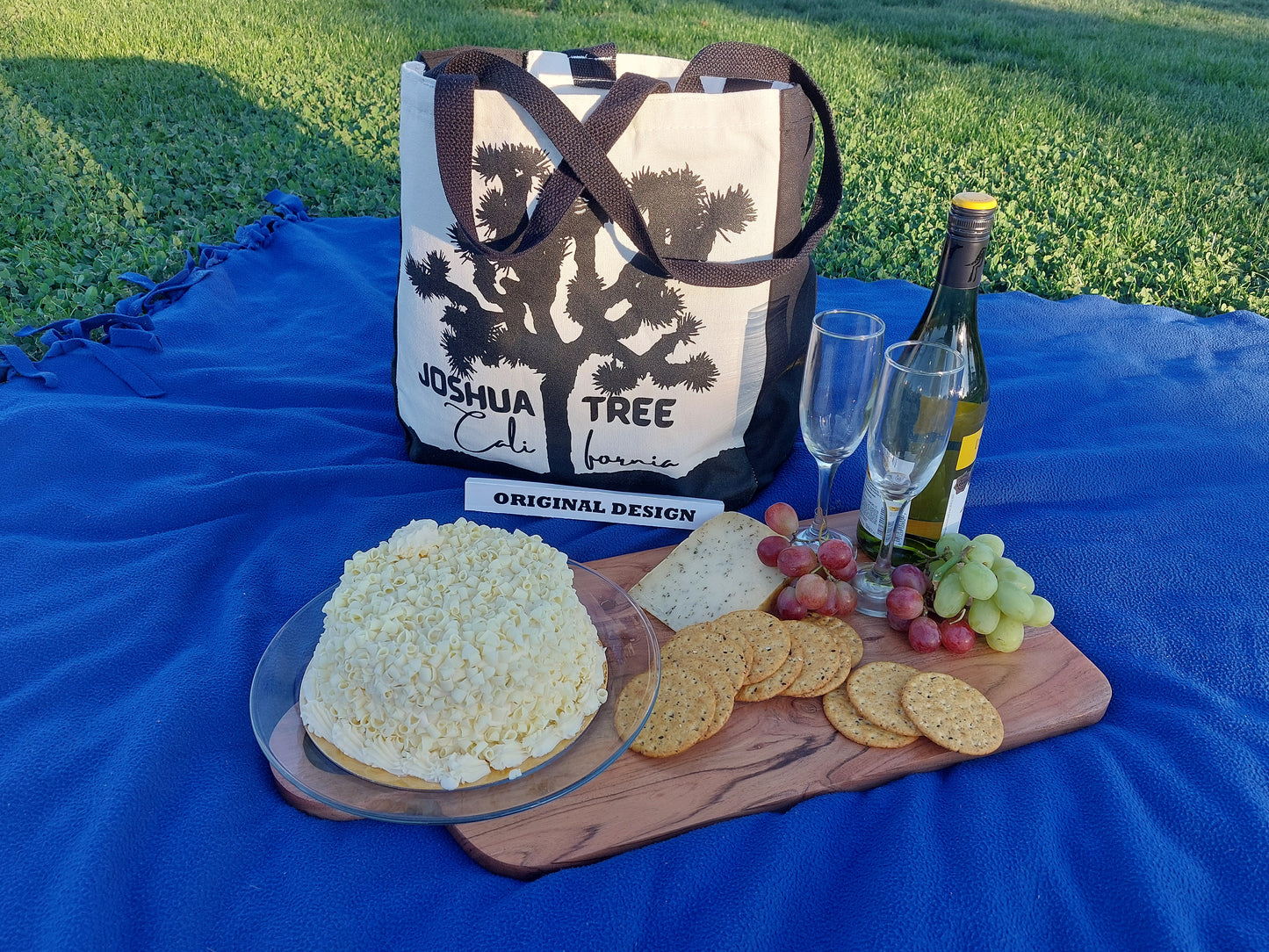 The image shows an ivory picnic bag with black handles. On the front of the bag is a black silhouette of a Joshua tree, with the words "JOSHUA TREE" and "CALIFORNIA" written below it. Our Picnic Bag is READY for ROMANCE! Large Enough to Carry A Full-Size Bottle of Champagne or Wine and Drinking Glasses as well as Food and a Picnic Blanket for those Last-minute Spontaneous GetAways!  
