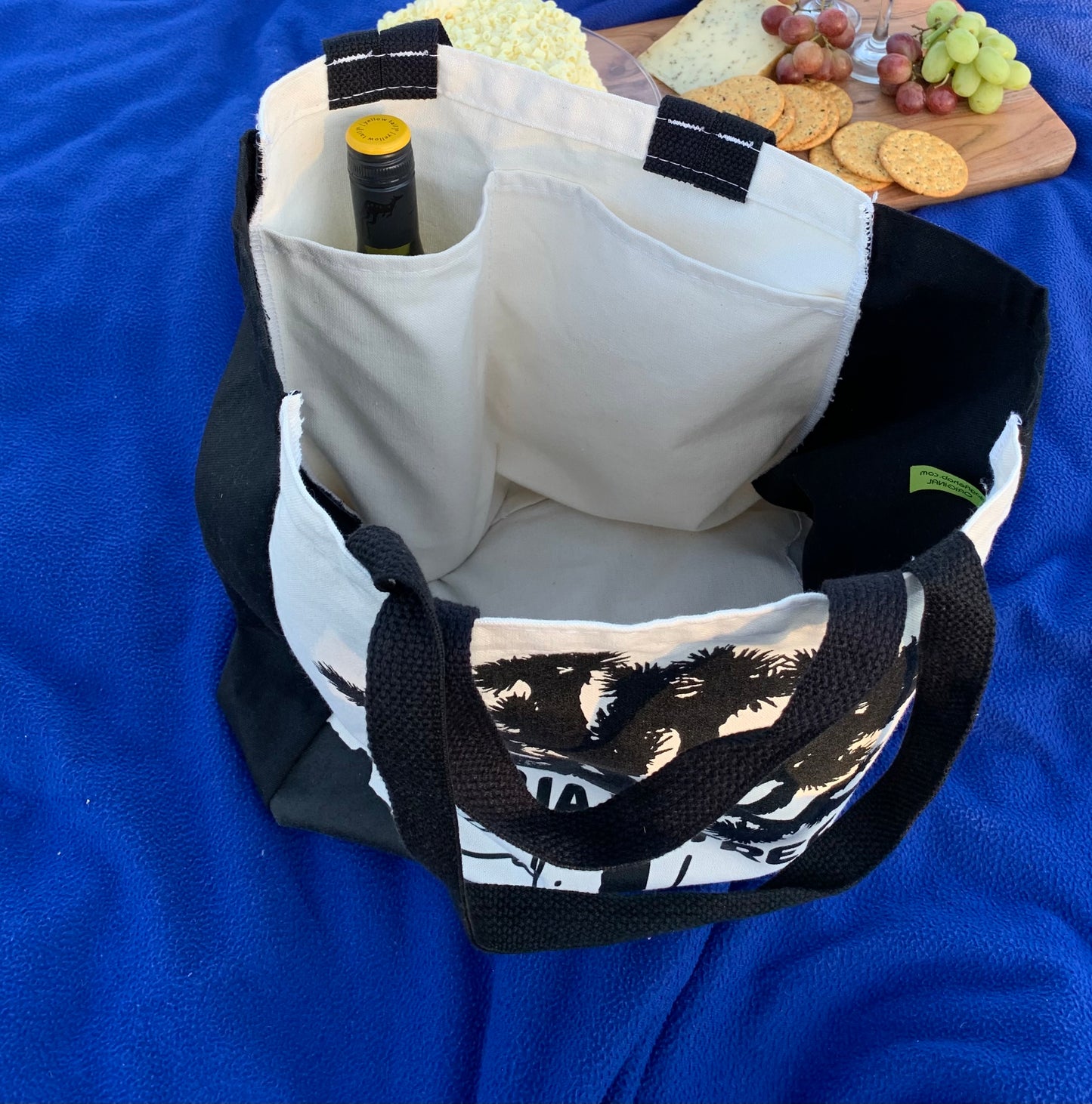 The image shows an ivory picnic bag with black handles. On the front of the bag is a black silhouette of a Joshua tree, with the words "JOSHUA TREE" and "CALIFORNIA" written below it. INSIDE are TWO Custom-designed Pockets to Hold a Wine Bottle or Champagne Bottle and Drinking Glasses as well as Picnic Food and Supplies and a Picnic Blanket.